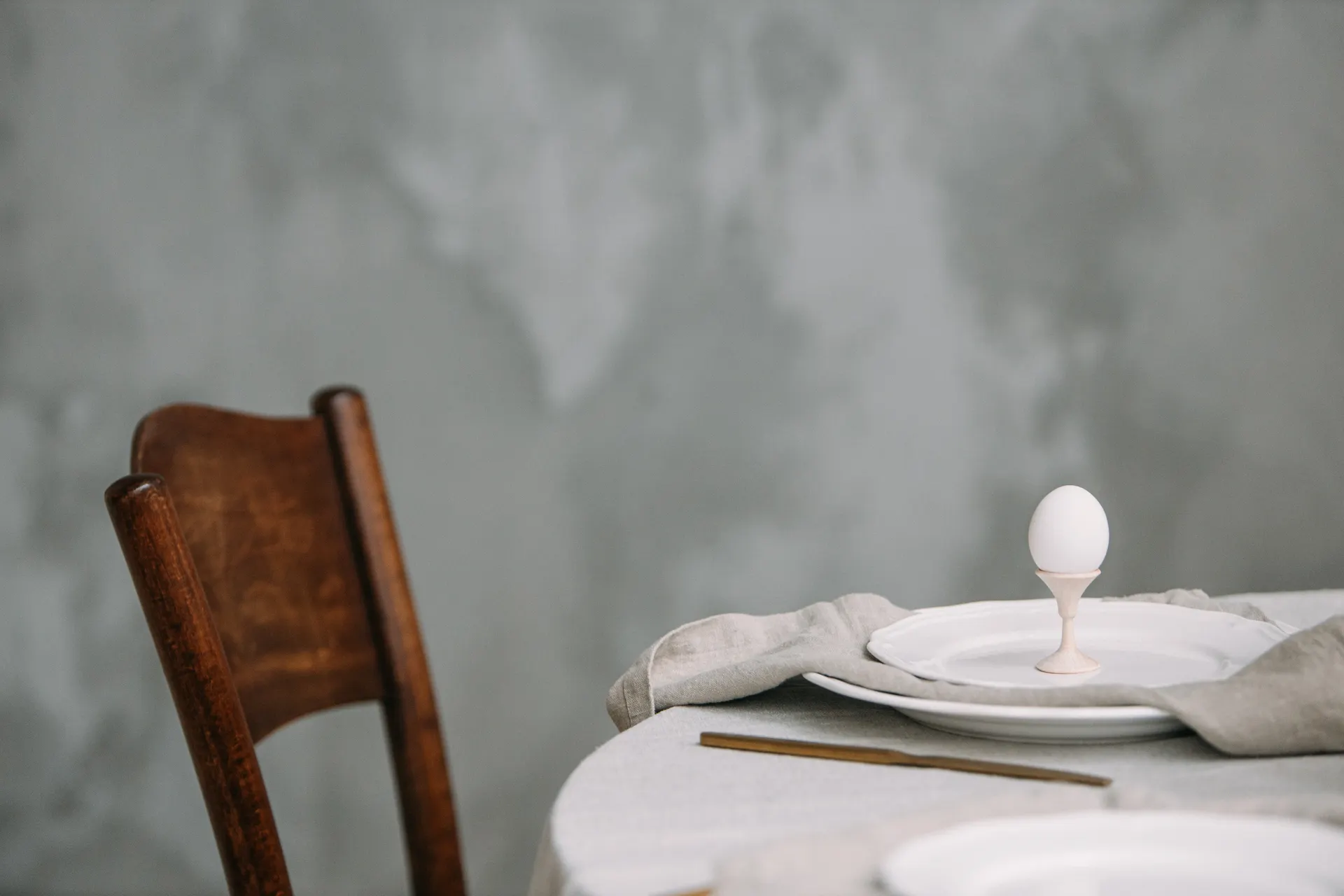 A quiet breakfast table: linen cloth, a wooden chair, an egg in a cup.